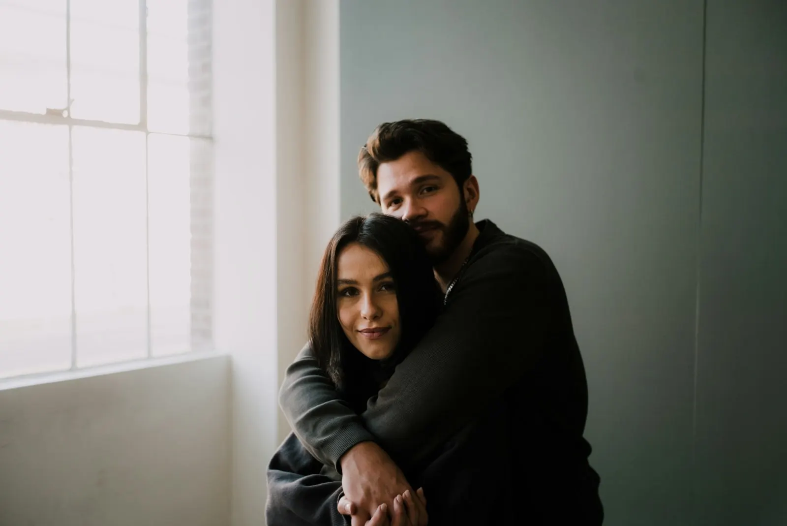 Pareja abrazada junto a una ventana: él la rodea con los brazos y ella mira tranquila